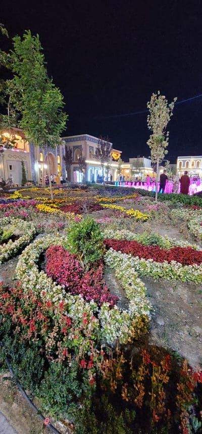 The image depicts a nighttime scene in a park or public square, possibly in the city of Satkak, Uzbekistan, as suggested by the prompt. The foreground is dominated by a large, elaborately designed flower bed with distinct sections of various colored flowers, including white, pink, red, and yellow. The flowers are arranged in curved rows and geometric patterns, creating a visually striking effect.
In the midground, several buildings with intricate architectural details are illuminated by artificial lights. These buildings appear to have traditional or ornate designs, with arched doorways and decorative tile work. People are visible walking around the buildings and in the background, suggesting a lively public space.
The background features a brightly lit fountain with colored water jets, adding to the festive atmosphere. Trees are scattered throughout the scene, some with lush green foliage and others appearing more sparse. The overall impression is of a well-maintained, beautifully decorated area, likely a popular spot for evening entertainment or relaxation. The lighting suggests it is after sunset, with the dark sky providing a contrast to the illuminated elements.