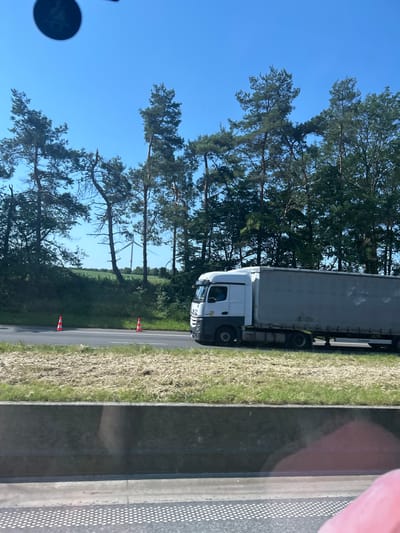 The image shows a white semi-trailer truck driving on a highway. The truck has a gray trailer. The highway is bordered by a concrete barrier and dry grass. In the background, there are trees and a wind turbine. The sky is clear and blue. There are no people visible in the image. The location is Jemeppe-sur-Sambre, Belgium.