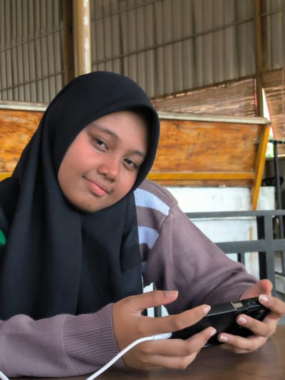 A person wearing a black hijab is seated at a table. The individual is holding a black smartphone, which is connected to a white charging cable. The person is wearing a long-sleeved top that is a muted purple color with a white and blue striped section on the left shoulder. The background includes wooden structures and a corrugated metal ceiling. Based on the location details, this scene is likely in Matang, Indonesia.