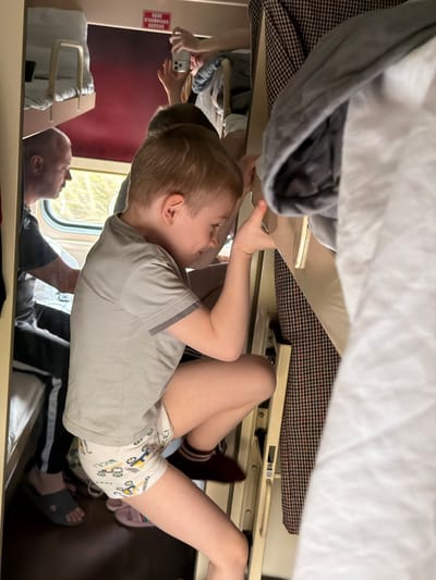 The image depicts a candid moment inside a train compartment, likely a sleeper car, set during the daytime in Bobrov, Russia. The main subject is a young, blonde-haired child, dressed in a light grey t-shirt with darker grey trim and white underwear shorts adorned with cartoon construction vehicles and the repeated word "NICE!". The child is positioned on hands and knees on what appears to be a lower bunk or seating area, leaning forward with a focused, curious expression, seemingly exploring or playing.

The scene is dominated by the train's interior, featuring bunk beds with white bedding in the foreground and a brown and black patterned fabric covering another surface. Natural light illuminates the compartment from a window on the right, through which indistinct greenery is visible, indicating the train is in motion.

Reflections on the ceiling and other polished surfaces provide additional details. An adult male's head is visible in reflection, looking towards the window. Another person is also reflected, holding a white smartphone (resembling an iPhone) pointed towards the child, suggesting they might be documenting the moment. These reflections also show parts of the train's interior, including a reddish wall and a beige handle or safety rail. Some white, illegible text is visible on a red panel within the reflection. Personal items such as slippers are scattered around, adding to the lived-in feel of the traveling environment. The overall atmosphere is one of domestic activity and travel.