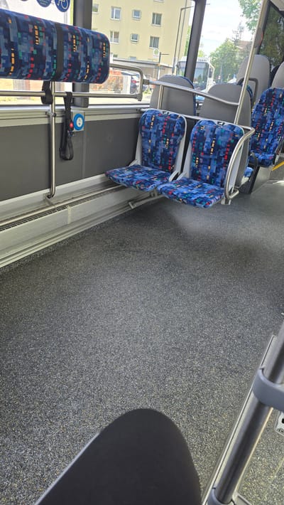 Inside a public transportation vehicle in Oberasbach, Germany, there are rows of blue patterned seats. A window with a view of a yellow building and trees can be seen. The floor is speckled gray and black. A black object is partially visible in the bottom right corner of the image.