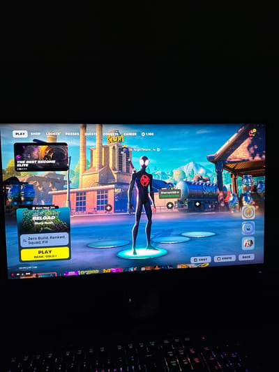 The image displays a computer monitor showing the lobby screen of a video game, likely Fortnite, rotated 90 degrees clockwise. A partially visible, backlit mechanical keyboard is to the right of the screen.

The game features a character resembling Spider-Man (Miles Morales suit) standing centrally on a circular platform. The background of the game is a detailed, bright, daytime environment with industrial-style buildings, barrels, and a blue sky.

Various user interface elements are visible on the game screen. Along the top, navigation tabs include "PLAY," "SHOP," "LOCKER," "PASSES," "QUESTS," "COMPETE," and "CAREER." On the far right, buttons for "CHAT," "EMOTE," and "BACK" are present. A player name, "19_Nigh7Mare_14," is displayed above the character, alongside an in-game currency count "1,100" (V-Bucks). Another player, "ShameAl186," is mentioned, with an option to "Reload Build Rani." The character's level, "LVL 16," is shown.

Game mode selections are prominent at the bottom left, including "THE BEST BECOME ELITE" and "RELOAD Slurp Rush" with a "Next Map 3m" timer. The active selection is "Zero Build, Ranked, Squad, Fill," with a large yellow "PLAY" button indicating "RANK: GOLD I." Performance metrics "120 FPS [1107 1120]" are visible at the very bottom left. Keyboard keys like "Alt + Shift" and function keys (F11, F12) are discernable on the physical keyboard.

This media file was captured in Bratya Daskalovi, Bulgaria.