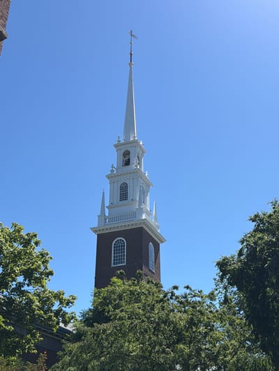 The image captures a prominent brick church steeple against a clear blue sky. The steeple is tall and features white architectural details, including decorative trim, arched windows, and a bell tower. A slender spire crowns the steeple, topped with a weather vane. Lush green trees frame the base of the steeple, partially obscuring a portion of the lower levels of the brick structure. The scene appears to be located in an urban environment, possibly a college campus given the architectural style and surrounding greenery. The location is the First Parish Church in Cambridge, Massachusetts, United States of America.