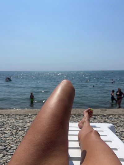 A first-person perspective shot captures a person's legs resting on a white lounge chair on a pebble beach. The person's right leg is in the foreground, with their foot visible at the bottom right of the frame. Their toenails are painted a bright green color. In the background, the calm sea stretches out under a clear blue sky. Sunlight glints off the water's surface. Several people are in the water, some swimming and others standing near the shore. The general atmosphere is one of relaxation and enjoyment of a sunny day at the beach. The location is the city of Blagoveshchenskaya, Russia.