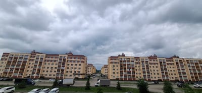 The image shows a street scene in Zubovo, Russia, dominated by a row of identical, light-colored apartment buildings with dark red trim and balconies. The buildings are multi-story structures with numerous windows. A gray, overcast sky hangs above the buildings. In the foreground, a paved road curves through the scene, with several vehicles visible. These include a dark blue truck in the left foreground, a white van further down the road, and several cars parked or driving on the street. There are also some trees and green areas alongside the road. The overall impression is of a residential area on a cloudy day.