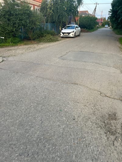The image shows a white car parked on the side of a paved road. The car is a modern sedan and has a license plate that appears to read "Y232BB 82". The road is surrounded by greenery, with trees and bushes on the left side of the frame and a house with a red tiled roof visible in the background. Further down the road, another white car is visible. The sky is overcast, suggesting it might be a cloudy day. The scene appears to be a residential area in Apsheronsk, Russia, possibly during daytime given the visible light. The image is taken from a slightly elevated angle, looking down the road. There are no people visible in the image. The overall impression is a quiet, everyday scene.