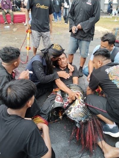 In Sumberejo, Indonesia, an outdoor scene depicts a group of individuals gathered around a man sitting on dark, wet ground. The central figure, a man wearing a black t-shirt and traditional black trousers with red and white patterns, appears fatigued or in an altered state, with a large, ornate black and white object resembling part of a traditional mask or costume with red and black fibrous elements resting over his lower legs. An individual wearing a black long-sleeved shirt, a dark vest, and a black headwrap with gold embroidery is embracing him from behind, with their head resting against his. Surrounding them are several other men: one stands to the left holding a decorated whip-like staff; another kneels to the right wearing a black t-shirt with a graphic design; a third sits to the left; and more figures are visible in the background crowd, near a red-draped structure on the left and a white building on the right. The ground is scattered with small, colorful debris.