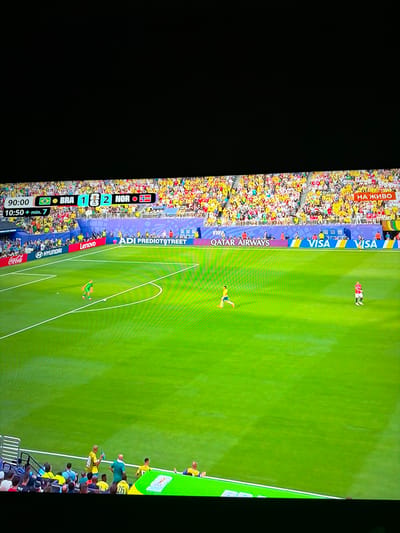 The image shows a soccer match being played on a bright green field under daylight. In the foreground, a player in a green uniform is running with the ball. Further down the field, another player in a red uniform is standing. The background is filled with spectators in a stadium, many wearing yellow clothing, cheering. A scoreboard at the top of the frame displays "90:00 BRA 1 2 NOR" and "10:50 + min. 7". Advertising banners for brands like Coca-Cola, Hyundai, Lenovo, Qatar Airways, and Visa are visible around the perimeter of the field. The overall scene depicts an active sports event. There is no indication of the location being Ognianovo, Bulgaria, within the image itself.