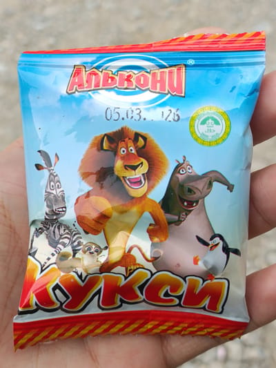 A close-up, eye-level, outdoor shot shows a hand holding a colorful, transparent plastic snack bag. The bag features characters from the animated movie "Madagascar," including Alex the lion, Marty the zebra, Gloria the hippopotamus, and Skipper the penguin. The text on the bag is in Cyrillic script, with "Альдони" at the top and "КУКСИ" at the bottom. A date "05.03.26" is visible. A green circular logo with Arabic writing, indicating Halal certification, is also present. The background appears to be a blurred outdoor environment with a rough, grey surface, possibly a street or pavement. There are no other people or significant features in the frame. The hand holding the bag is partially visible on the right and bottom edges.