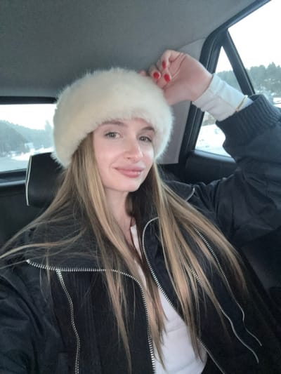 A single female subject, appearing to be an adult, is seated in the rear passenger compartment of a vehicle in Razvršje, Montenegro. Her long, straight, light brown hair with blonde highlights is visible beneath a cream-colored, fluffy, round winter hat. She is wearing a black puffer jacket with prominent silver zippers and a white long-sleeved shirt. A thin, gold-toned bracelet is present on her left wrist. Her left hand is raised near her head, fingers slightly curled, displaying red-painted fingernails. The subject is looking directly forward with a subtle, closed-lip smile. The vehicle's interior features a black headrest and dark trim. Through the window to the right, an outdoor scene depicting snow-covered ground, dark coniferous trees, and an overcast sky is visible, indicative of a winter landscape.