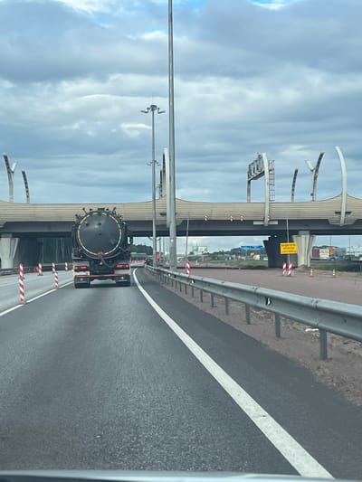 A black, circular tank truck is driving on a highway. The truck has a large, black cylindrical tank with yellow and white stripes around its circumference. It has multiple pipes and valves visible on the back of the tank. The truck is in the right lane of a multi-lane highway. In the distance, an overpass spans the highway, with large concrete pillars supporting it. The sky is overcast with gray clouds. The scene appears to be on the outskirts of a city, with vegetation visible beyond the highway on the left. There are orange traffic cones and striped traffic barriers along the side of the road. A yellow sign with black text is visible under the overpass.