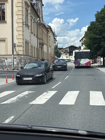 In Zell am See, Austria, on a clear day with scattered clouds, a black Audi A4 Avant is stopped at a pedestrian crossing on a street lined with traditional European architecture. The car, bearing the license plate "ZE 745 IJ", is positioned in the right lane. Ahead of the Audi, a black Skoda SUV is also stopped, its license plate reading "1A1 8733". Further down the street, a multi-colored bus with "Salzburg Verkehr" branding is in motion, also approaching the pedestrian crossing. Two young males, dressed casually in t-shirts and shorts, are walking on the sidewalk to the left of the Audi, one of them carrying what appears to be a white object. Several other pedestrians are visible further down the street, near the buildings. The street is paved and marked with white lines for the pedestrian crossing and traffic lanes. Orange traffic cones are placed on the side of the road. The buildings feature stone facades and tiled roofs, consistent with Austrian urban design. The overall scene depicts a typical day in a European town, with vehicles and pedestrians navigating a public street.