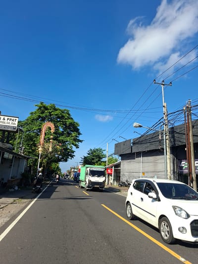 This image depicts a street scene in Banjarangkan, Indonesia, under a clear blue sky with scattered white clouds. The road is asphalt, with a yellow line marking the center lane and a white line bordering the right side. On the left side of the road, there is a sidewalk and buildings, including one with a sign that reads "S & PPAT MAYASA, S.H., M.Kn.". A large green tree stands prominently behind a decorative archway.
Several vehicles are present: a white car is in the foreground on the right, a green Isuzu truck is in the middle of the road, and several motorcycles are visible further down the street. People are seen riding on the motorcycles. Electrical poles with numerous wires crisscross the sky above the buildings. The overall environment suggests a busy urban or semi-urban area.