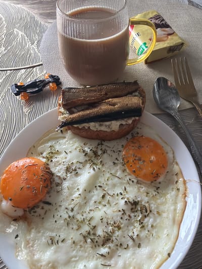 The image displays a breakfast setting on a textured, possibly wooden, surface. A white plate holds two fried eggs, one with a visible yolk, and a half of a bun topped with what appears to be a spread and several smoked sprats. The eggs and bun are seasoned with dried herbs. To the right of the plate, a fork and spoon are visible. A glass mug filled with a light brown beverage, likely coffee with milk, sits further back. Beside the mug is a yellow package of cookies. In the upper left corner, a small, dark toy car with orange wheels is positioned. The overall scene suggests a meal being prepared or consumed indoors.