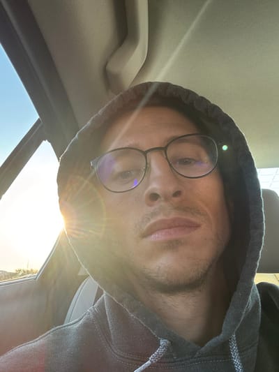An image depicts a male individual, appearing to be in his 20s or 30s, with light stubble, wearing dark-rimmed eyeglasses and a dark gray hooded sweatshirt with the hood pulled over his head. He is positioned in the passenger seat of a vehicle, looking directly at the camera with a neutral expression. Strong, low-angle natural sunlight from the left side of the frame creates significant lens flare, partially obscuring the left side of his face and the vehicle's exterior view. A clear blue sky is visible through the vehicle's window on the left. The vehicle's light-colored interior headliner is visible in the upper portion of the frame. The scene is located within the geographical area of Ramat Negev Regional Council, Israel.