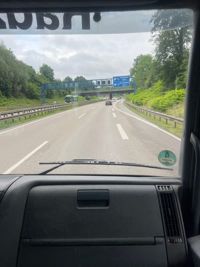 The image is taken from the interior of a vehicle, likely a truck, looking forward through the windshield. The viewpoint is from the driver's seat. The road ahead is a multi-lane highway, seemingly an Autobahn, with vehicles in motion. Overhead signs indicate directions to "Bö-Eppendorf 1200 m" and "Bö-Stahlhausen 600 m." The terrain to the right of the highway is a steep, green embankment covered in dense foliage. On the left side, a metal guardrail lines the road, with trees and greenery beyond. The sky is overcast with clouds. A circular sticker with green and white imagery is visible on the lower right portion of the windshield. The dashboard of the vehicle is visible in the foreground, showing the steering wheel area and a ventilation grille. The lettering on the top of the windshield appears to be part of the vehicle's branding or registration, but is largely obscured and illegible. Based on the highway signage referencing "Bö-Eppendorf" and "Bö-Stahlhausen," which are districts within Bochum, Germany, this scene is situated on a highway in or near the city of Bochum.