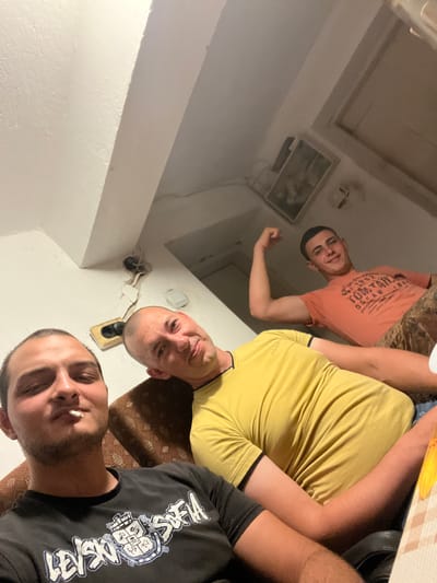 The image shows three adult males in an indoor setting. Two of the men are in the foreground, one on the left with a cigarette in his mouth, and another in the center, looking towards the camera with a slight smile. A third male is in the background, flexing his bicep and smiling. They appear to be in a living space, with a couch and a wall with exposed electrical wiring and an electrical outlet visible. The lighting is dim, suggesting an indoor environment. The location is identified as Dragoevo, Bulgaria.