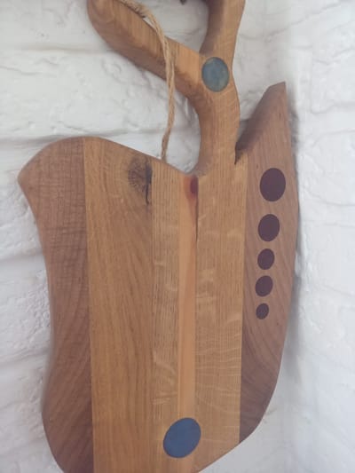 The image displays a wooden cutting board with a unique, carved handle hanging against a white brick wall. The cutting board is made of several different wood planks joined together, showcasing variations in grain and color. It features decorative circular inlays of dark purple and blue. A rough-hewn jute rope is attached to the handle for hanging. The overall scene is indoors, with the lighting suggesting a natural light source from the side. There are no people present in the image. The context of this object being located in Terebovlia, Ukraine, is not visually ascertainable from the image itself.