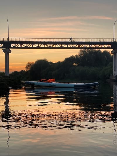 A wide-angle, eye-level shot captures a serene sunset scene over a body of water, likely the Berezina River in Barysaw, Belarus. The sky displays soft hues of orange, pink, and pale blue, with wispy clouds. A long, modern bridge with a metal truss structure stretches across the frame, supported by concrete pillars. A solitary cyclist is silhouetted against the sky, riding across the bridge. Below the bridge, dense green trees line the riverbank. In the foreground, a blue and white paddleboard with an orange life vest is floating on the calm water. The water's surface reflects the warm colors of the sunset and the dark shapes of the trees and bridge, creating a mirror-like effect.