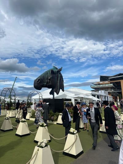 The image depicts a daytime outdoor scene at the Ascot Racecourse in Ascot, United Kingdom. The sky is partly cloudy with a mix of grey and white clouds against a blue sky. In the foreground, a large, dark bronze sculpture of a horse's head is prominently displayed. The sculpture is weathered, with a greenish patina visible.

Surrounding the sculpture is a crowd of people dressed in formal attire, many wearing hats, indicative of a prestigious event like Royal Ascot. The ground is covered with artificial green turf, and white pyramidal planters filled with white flowers are arranged along pathways, demarcated by ropes.

In the midground, several white marquees or tents are set up, suggesting hospitality or event areas. Behind these tents, a modern multi-story building with balconies and white awnings, which is characteristic of the Ascot Racecourse grandstands, is visible. To the left of the frame, a green construction crane is present, hinting at ongoing development or maintenance.

The people in the image are walking and standing, some are engaged in conversation. The overall atmosphere appears to be one of a social gathering at a sporting event. The focus is on the sculpture, the people, and the architectural elements of the racecourse.