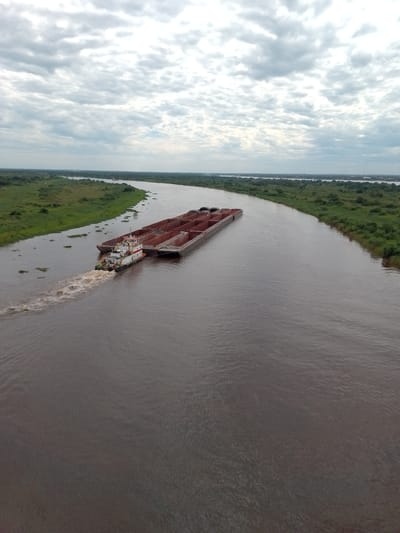 A long, wide river winds through a green, flat landscape under an overcast sky. A tugboat pushes a large barge loaded with rectangular containers up the river. The tugboat leaves a trail of churned water behind it. The riverbanks are covered in dense vegetation. The image is taken from a high vantage point. The location is Concepción, Paraguay.