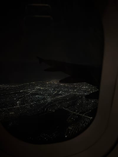 The image is an aerial view through an airplane window at night, looking down upon a sprawling urban landscape illuminated by countless lights. The lights form a dense, interconnected network across the dark terrain, suggesting a significant population center. The wing of the airplane is visible in the foreground, partially obscuring the view of the city below. The city's lights are blurred, indicating motion or a long exposure time. The overall impression is one of a vast, illuminated city at rest under the darkness of night, seen from a high vantage point. The location is not identifiable from the image alone and no specific details about Асқар Тоқпанов ауылы can be extracted.