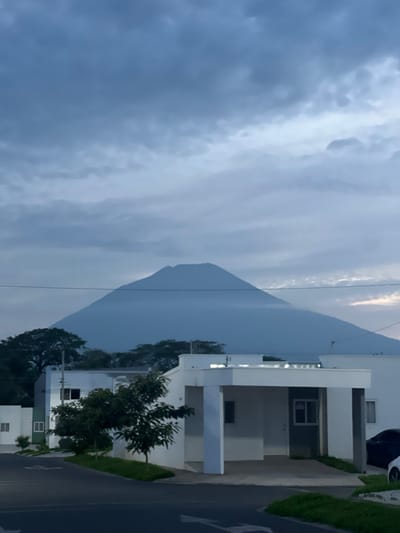 The image depicts a serene, possibly pre-dawn or dusk, outdoor scene with a prominent volcano in the background, characteristic of the region surrounding San Miguel, El Salvador. In the foreground, modern, white-and-gray residential buildings with flat roofs and minimalist architectural designs are visible. The houses appear to be part of a planned community, with well-maintained landscaping featuring small trees and green grass. A paved road curves through the foreground, with white directional arrows painted on its surface. The sky is overcast with soft, diffused light, casting a slightly muted tone over the landscape. No people or vehicles are clearly discernible, contributing to the peaceful and quiet atmosphere of the scene. The overall impression is one of modern residential living in a picturesque natural setting, with the imposing volcano serving as a constant, majestic backdrop.