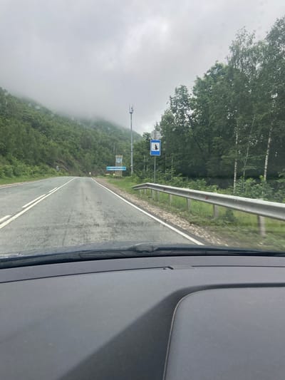 The image captures a view from inside a vehicle traveling on a paved road through a mountainous, forested area. The sky is overcast and foggy, obscuring the tops of the distant hills. A communication tower with antennas is visible on the right side of the road. Further down the road, a blue sign with a white icon of a pine tree is displayed. On the right, a metal guardrail runs alongside the road, separating it from a grassy embankment and trees. On the left, the road curves slightly and is bordered by a steep, tree-covered mountainside. Two sets of white lines mark the lanes of the road. There are no people or animals visible in the scene. The dashboard of the vehicle is visible in the foreground. The scene appears to be on a road leading towards or from Usakly, Russia, a town known for its natural beauty and mountainous terrain.