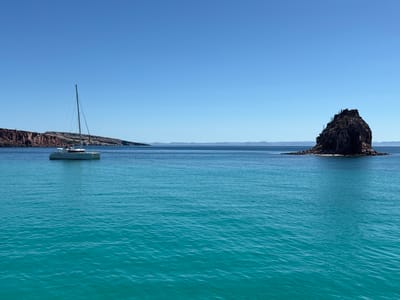 The image presents a coastal scene in what is described to be the city of Unknown, Mexico. The scene features a clear blue sky above a tranquil body of water, a turquoise sea that extends to the horizon. A white catamaran sailboat is positioned on the left side of the frame, anchored or drifting near the shoreline. A rocky islet, capped with sparse vegetation or cacti, stands prominently to the right. In the background, a mountainous coastline can be discerned. The overall impression is one of serenity and natural beauty.