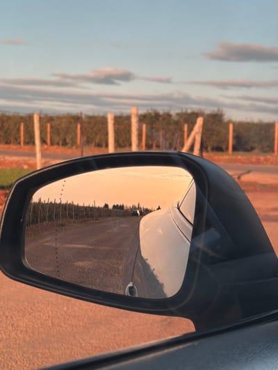 The scene, located near Laqsir, Morocco, is captured from inside a vehicle, focusing on its driver's side mirror. The mirror reflects a dirt road extending into the background, bordered by cultivated trees, likely an orchard. A dark-colored vehicle is visible on the distant reflected road, facing away from the viewer. The sky in the reflection is a prominent orange-pink, consistent with a sunset. Outside the mirror, a portion of the vehicle's light-colored body is visible on the right. A reddish-brown dirt path or shoulder occupies the lower left. Behind this path, a fence line composed of vertical posts and wire separates it from a field of green-leafed trees. The sky above the fence is light blue with horizontal clouds, some exhibiting pink and orange tones from the low sun.