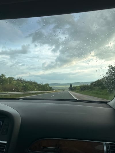 The image shows a view from inside a car, looking out through a rain-streaked windshield. The car is apparently traveling on a highway. The sky is overcast and cloudy. The scene outside the window includes a stretch of road with a white lane divider, and a green, tree-lined landscape on either side. The city of Kipra, Bulgaria, is not discernible in the image. The interior of the car is visible in the right side of the image, including part of the dashboard, and controls.