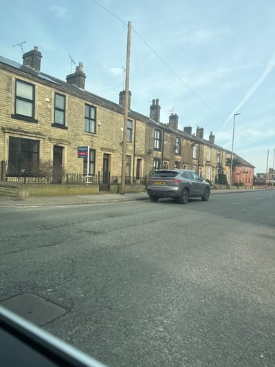This image depicts a residential street in Bury, United Kingdom, observed under a bright, clear sky with a visible contrail. The foreground features an asphalt road surface with noticeable cracks and a utility access cover in the lower left. A modern grey SUV, registered "MJ74 NWO", is parked curbside on the left, facing away from the viewer.
To the left, a continuous row of two-story terraced houses, constructed from light-colored stone, lines the street. These houses feature dark-framed windows, dark front doors, and chimneys on their roofs; solar panels are visible on the roof of the leftmost house. Small front gardens are delineated by low stone walls and black wrought-iron railings and gates. A red and white "Harrison SOLD" sign is prominently displayed in the garden of house number 65, located centrally among the visible houses. A wooden utility pole with overhead wires runs along the left side of the street.
The street recedes into the distance, revealing additional terraced houses, some with reddish-painted facades, on both sides. A streetlight is visible on the right side of the road further down. The scene is devoid of people.