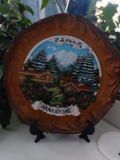 A decorative plate, likely a souvenir, is displayed on a white surface, possibly a shelf or windowsill, with a dark stand supporting its base. The plate features a raised relief depicting a winter mountain scene. Snow-capped mountains rise in the background under a pale blue sky with some white cloud-like formations. In the mid-ground, several snow-laden evergreen trees are prominent, and nestled among them are two small, rustic wooden houses with red roofs, illuminated by warm, yellow light from their windows. The foreground of the plate is adorned with lush green and pink foliage. A white banner across the bottom of the scene displays text in Cyrillic script, which reads "ЭЛЬБРУС" (Elbrus). Above the mountain range, the numbers "5642" are visible, likely representing an elevation. The plate itself has an ornate, dark brown, carved frame with a scalloped edge. To the left and right of the plate, out of focus, are green leaves of a plant, suggesting it might be near a houseplant. The lighting suggests daylight, with a bright, possibly sunlit, area visible in the upper right corner, creating highlights on the plant's leaves. There are no people or animals depicted in the image. The location is not identifiable as Novopokrovka, Kyrgyzstan, based on the visual information presented.