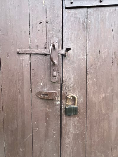 The image shows a weathered, dark brown wooden door with vertical planks. A metal latch mechanism with a handle is visible on the left side of the door. A hasp is attached to the door, and a green padlock is secured to it, indicating the door is locked. The wood appears old and worn, with some paint peeling and visible grain. The environment suggests an outdoor setting, potentially in the rural area of Бучацінскі сельскі Савет, Belarus, given the rustic nature of the door and its hardware. There are no people or animals present in the image.