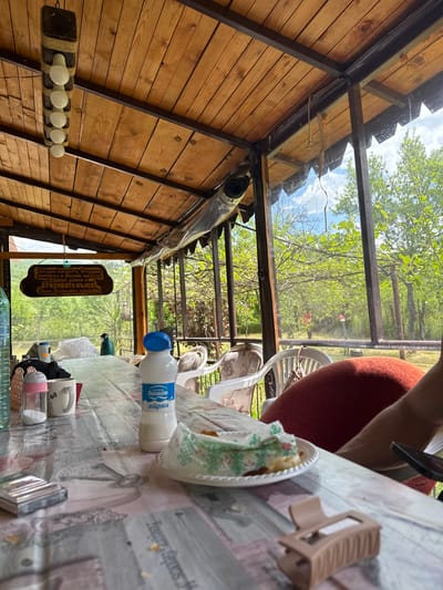 The image shows an interior view of a structure, possibly a gazebo or a summer house, with a wooden frame and a roof that allows a view of the outside, featuring trees and sky. Inside, a table covered with a patterned tablecloth is laden with various items: a plate of food, a bottle of Balkan Ayran (a yogurt-based drink), mugs, a baby bottle, and other objects. A sign indicating the location as "Розовата къща" (The Pink House) in the village of Praveshka Lakavitsa, Bulgaria, hangs near the structure's frame. A person is partially visible near the table, suggesting the scene is set during a meal or gathering. The overall atmosphere seems relaxed, with the outdoors visible and a meal being enjoyed inside the structure.