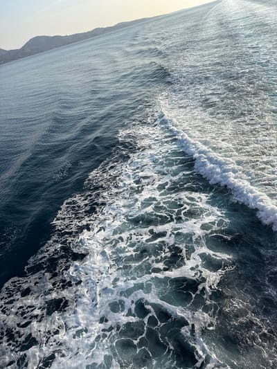 The image presents an angled, high-level view of the Aegean Sea near Kamariotissa, Greece. The scene is dominated by a turbulent expanse of water, specifically a white, aerated wake. This wake forms a distinct diagonal line from the bottom right towards the upper center, separating calmer, deep blue water on the left from a wider zone of foamy, light blue-green water on the right. A prominent breaking wave crest is visible within the turbulent zone. In the upper left quadrant, a segment of coastline belonging to Samothrace island is discernible, characterized by gently sloping hills under a bright, hazy sky. No individuals or vessels are visible in the frame, though the presence of a wake implies a moving watercraft just outside the view.