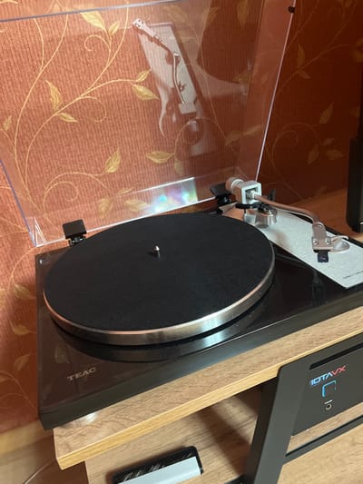 The image shows a close-up, eye-level view of a black TEAC direct drive turntable. The turntable has a silver trim around its black platter, which has a textured surface. A tonearm with a silver finish is positioned above the platter. The turntable is placed on a light wood shelf unit. In the background, there is an orange textured wallpaper with a subtle gold floral pattern. To the right of the turntable, a black electronic device with a blue illuminated display that reads "IOTAVX" is visible. The scene appears to be indoors, likely in a domestic setting. There are no people or animals present in the image, and no specific location in the city of Яново-Грушевский, Russia can be identified from the visual information provided.
