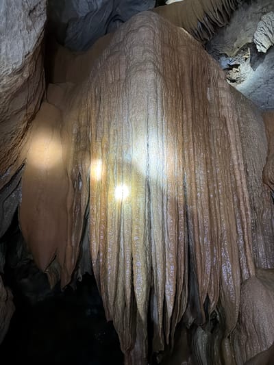 The scene depicts the interior of a natural cave in Phang-nga, Thailand. A large, prominent geological formation, identified as a wet, reddish-brown to tan flowstone or drapery, dominates the central and right portions of the frame. This formation displays extensive vertical corrugations and folds, with a glistening surface indicating the presence of moisture. Multiple localized, intense bright spots, consistent with artificial light sources such as flashlights, illuminate the wet rock, creating distinct reflections. Surrounding rock walls and ceilings, characterized by rough textures and darker tones, are visible in the subdued ambient lighting of the cave. No individuals or specific objects of human origin are visible within the frame, though the presence of artificial illumination implies human activity or exploration within the cave.