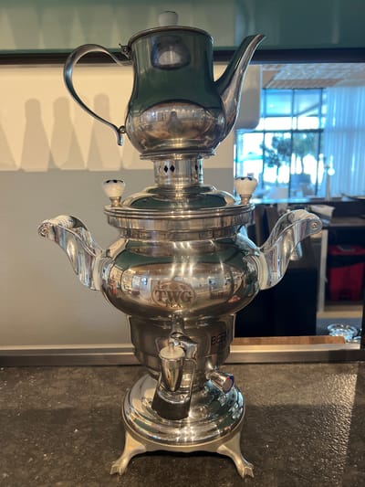 This is a close-up, vertically oriented photograph of a highly polished, silver-colored samovar. The samovar is the primary subject, with its intricate metalwork and reflective surface dominating the frame. It features a spout, handles, and decorative elements, including what appears to be a circular logo with text. The samovar is positioned in what seems to be an indoor setting, possibly a kitchen or dining area, with a dark, textured countertop visible in the lower right portion of the image. The background shows a window with blinds and diffused natural light, suggesting it might be daytime. There's also a hint of shelving and some red objects in the blurred background.
The samovar has a prominent spout on the left and an ornate handle arching over its body. A smaller handle or knob with blue and white detailing is visible on the side. The reflection on the samovar's surface is significant, showing distorted images of the surroundings, including the window and possibly other objects not clearly identifiable. Text is visible on the samovar, including "BEEM" and within the circular logo, "TWO TEA". The overall impression is one of a decorative and possibly functional kitchen appliance, captured in a well-lit environment. The image does not depict any people or specific weather conditions, but the lighting implies daytime. The location context suggests it is in Ras Al-Khaimah, United Arab Emirates.