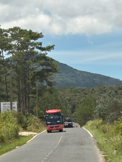 The image depicts a paved road winding through a landscape characterized by dense green trees and rolling hills under a partly cloudy sky. A bright red bus is in the foreground, traveling away from the camera. To the left of the road, there is a white sign with text, partially obscured by foliage. Further down the road, a dark-colored van and a person on a scooter are visible in the distance. The road is lined with grass and small red and white markers. The overall scene suggests a rural or mountainous area. While the prompt requests information about the location in the city of Đưng K'Si, Vietnam, this specific detail cannot be confirmed solely from the visual information in the image.