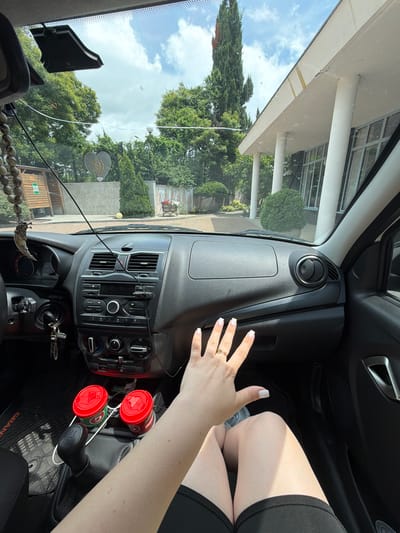 The image shows the interior of a car, looking outwards through the windshield. The scene is bright, suggesting daytime. The driver's hand, with manicured nails and a ring, is visible in the foreground, resting on the dashboard. Two red-capped cylindrical objects, possibly drink containers, are placed in the center console. Outside, lush green trees and a partly cloudy sky are visible, indicating fair weather. A building with white pillars is also present in the background, suggesting a residential or public area. The car's dashboard features air vents, a radio, and various control knobs. A string of beads hangs from the rearview mirror. The words "GRAND" are partially visible on the car mat. The scene appears to be a snapshot of a moment during a car journey on a pleasant day.