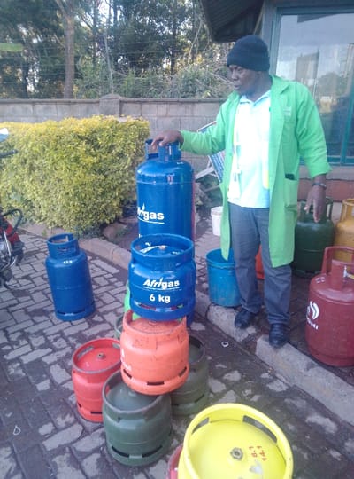 The image depicts an outdoor commercial scene in Karai ward, Kenya, featuring a male individual amidst numerous liquefied petroleum gas (LPG) cylinders. The ground is paved with wet, interlocking grey concrete bricks.
A male of African descent stands on the right, wearing a black knitted beanie, a light blue short-sleeved polo shirt, dark grey trousers, black laced shoes, and a light green long-sleeved overcoat with "BIGGAS" visible on the left chest. His right hand rests on the handle of a tall, blue "Afrigasi" branded LPG cylinder. He is looking towards the left side of the frame.
Approximately twelve to fifteen LPG cylinders of various sizes and colors are arranged across the paved area. Blue cylinders include a tall "Afrigasi" unit, a smaller "Afrigasi 6 kg" unit stacked on an orange cylinder, and another blue cylinder marked "12.0" further left. Other cylinders are orange, green, red (one with a partial "Hashi" logo), and yellow, many stacked.
In the background, a low stone wall is topped with a fence or wire, behind which dense green foliage forms a hedge, and tall trees are visible. To the far right, a building with a dark roof overhang and a large window is partially visible. A white plastic bucket and a blue plastic bucket are positioned near the man's feet by the building. On the far left, the front wheel and handlebars of a red motorcycle are partially visible.