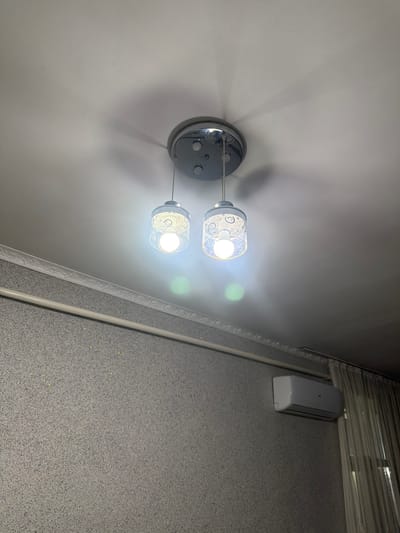 The image shows a close-up view of a ceiling light fixture with two glass pendant lamps, each containing a bright LED bulb. The lamps have a decorative pattern of swirls. A shadow from the light fixture is cast on the ceiling. Below the ceiling, there is textured wallpaper with a decorative molding strip running horizontally. A white air conditioning unit is mounted on the wall, and beige curtains are visible to the right. The overall scene suggests an indoor residential setting, possibly in Aleksandrovka, Kyrgyzstan.