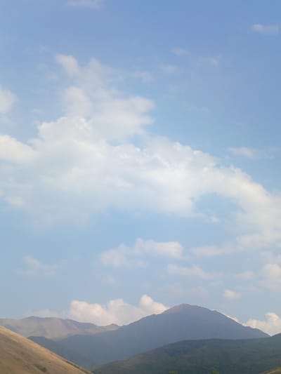The image depicts a mountainous landscape under a partly cloudy sky. In the foreground, a sloping hillside, rendered in earthy tones, leads the eye towards layers of distant mountain ranges. The peaks are softened by atmospheric haze, with varying shades of purple and blue indicating depth. The sky is a pale blue, punctuated by scattered white clouds of varying sizes and densities. The overall impression is one of natural tranquility and vastness, characteristic of high-altitude environments. The scene is devoid of human presence or man-made structures.