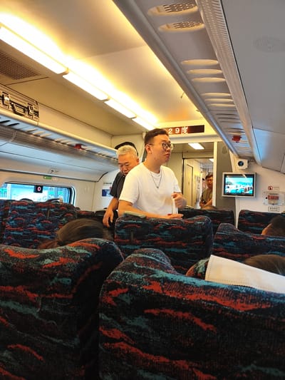The image captures the interior of a train carriage, likely traveling through or to Harbin, China, given the presence of the Chinese characters "哈尔滨" (Harbin) on a sign visible in the background. The scene depicts several passengers seated on colorful patterned upholstery. In the foreground, the back of a person's head with dark hair is visible. A man in a white t-shirt and glasses is standing, looking to his left, and holding a white object. To his left, another man with gray hair, wearing a black shirt, is also present. The train carriage features overhead lighting, luggage racks with bags, and a small screen displaying a scenic image. A temperature display showing "29度" (29 degrees) is mounted above the aisle. The overall impression is of a typical public transportation environment.