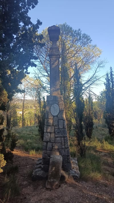 The image shows a stone monument located in a grassy, wooded area in Jérica, Spain. The monument consists of a stacked stone base supporting a tall, narrow stone pillar topped with an ornate, urn-like structure. A circular plaque is embedded in the stone base. The monument is framed by trees and shrubs, with a landscape visible in the background under a blue sky. The ground is covered with grass and dirt. No people are visible.