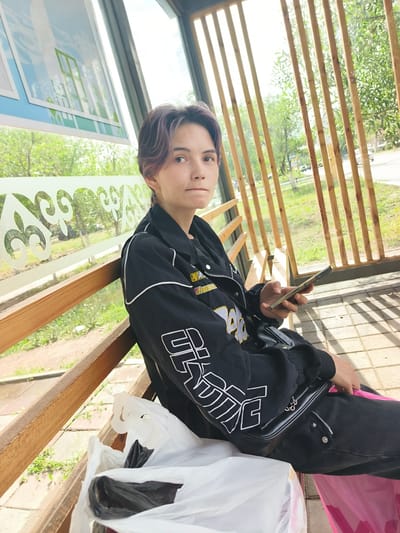 A young person with short, layered, purplish-brown hair sits on a wooden bench at a bus stop in Arkalyk, Kazakhstan. They are wearing a black bomber jacket with white trim and large white lettering on the sleeves, black pants, and a small black purse with a silver chain strap. They are holding a smartphone in their right hand.

In front of them are two plastic bags, one white and one black, and a pink shopping bag. The bus stop has a wooden bench and a roof supported by wooden beams. The background shows green trees and a street, suggesting it is daytime and likely spring or summer. The overall atmosphere is casual and relaxed, with the person appearing calm as they wait. There is a decorative patterned glass panel along the side of the bus stop. The weather appears to be clear and sunny.