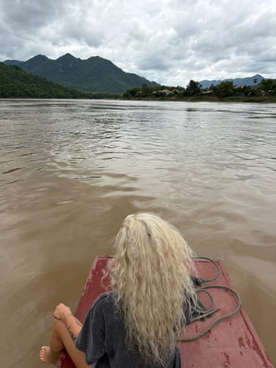 A person with long, blonde, curly hair is sitting on the front of a red boat, facing away from the camera, looking out at a wide, muddy river. The person is wearing a dark gray t-shirt and has tattoos on their arm and leg. Their legs are crossed, with their right knee bent and their left leg extended. A rope is coiled on the boat near the person. The river is calm with ripples on the surface, reflecting the overcast sky. In the background, lush green mountains rise up on either side of the river, with a small village visible on the right bank. The sky is heavily clouded with gray tones, suggesting a cloudy or possibly rainy day. The scene appears to be located in or near Houaymat, Laos.