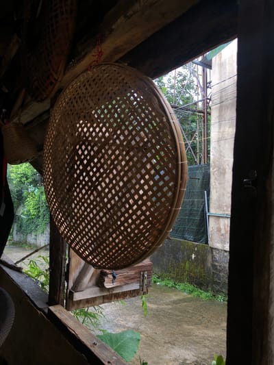 A low-angle, medium shot captures a woven bamboo basket hanging in a rustic wooden structure, possibly a veranda or an open-air room. The basket, intricately woven with a diamond pattern, is the primary subject, occupying the center-left of the frame. It appears to be an outdoor setting in the city of B'Lao, Vietnam, as indicated by the dense green foliage, a concrete wall with moss growth, and power lines visible in the background. The lighting is dim, suggesting it is either early morning or late afternoon. The basket is suspended by a thin red string from a weathered wooden beam above. To the left of the basket, another smaller woven basket is partially visible, hanging lower. The foreground is dominated by rough, aged wooden planks, forming part of the structure the baskets are hanging from. The background reveals a wet, possibly rain-slicked, outdoor area with scattered green plants and a hint of a building. The overall scene evokes a sense of rural or traditional Vietnamese life, with an emphasis on natural materials and a slightly unkempt, lived-in aesthetic. There are no people or animals in the frame.