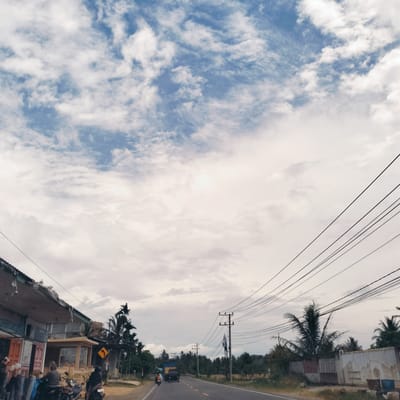 This media file captures a wide-angle view of a roadside scene in Pandrahkandeh, Indonesia. The setting depicts a relatively quiet day along a two-lane asphalt road with a yellow double solid line.
On the left side of the road, several single-story buildings, likely shops or homes, are visible. Their roofs appear somewhat worn or corrugated. In front of these buildings, several motorcycles are parked or stopped, and a few individuals are present, including one person wearing a beanie and another wearing a helmet. A yellow diamond-shaped road sign with a black arrow indicating a right curve is prominently displayed near the buildings.
Further down the road, a blue and black truck or bus is visible, driving away from the viewer. On both sides of the road, particularly in the mid-ground and background, dense tropical vegetation, primarily palm trees, flourishes. Numerous wooden utility poles with a complex network of overhead power lines crisscross the sky, extending diagonally across the frame, especially on the right side. A white and brick fence or wall is also seen on the right, further down the road.
The sky is largely covered with clouds, ranging from patchy white clouds to a more diffused overcast layer, with glimpses of blue sky showing through. The overall lighting suggests it is daytime, possibly late morning or early afternoon, with no strong shadows indicating direct sunlight. No discernible text is visible on any signs or objects. The scene conveys a sense of everyday life in a rural or semi-urban Indonesian community.